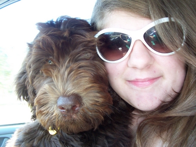 A woman wearing white sunglasses takes a selfie with a brown shaggy dog.